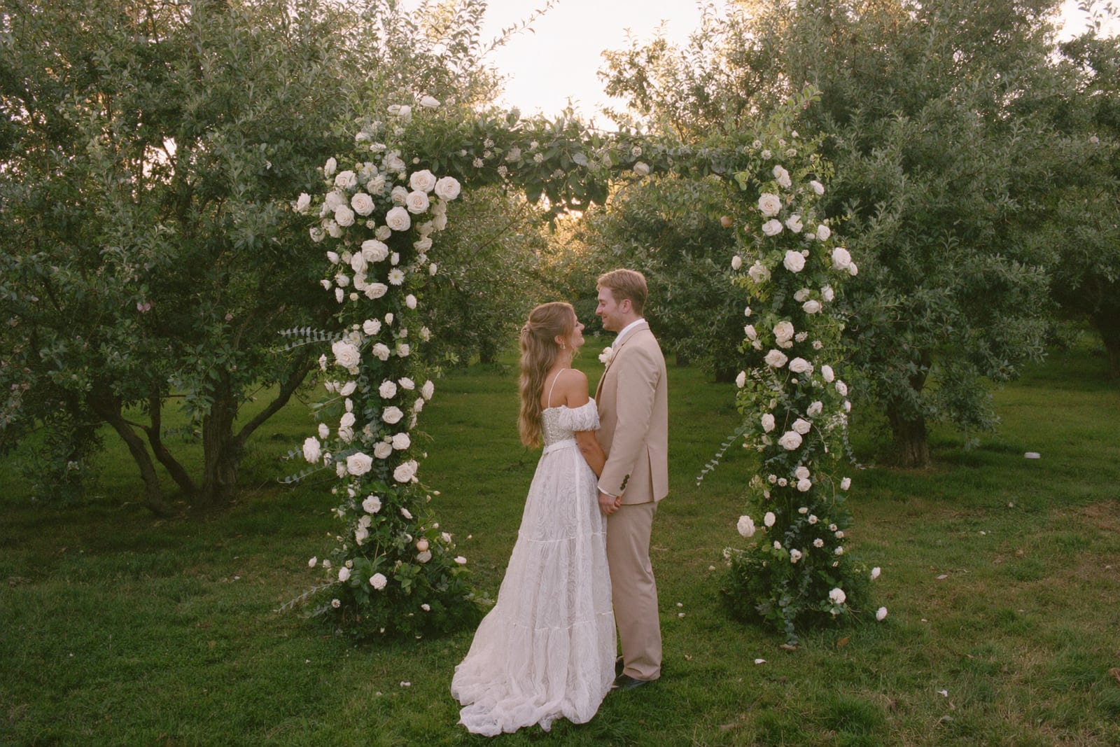 Elise en Thomas hand in hand onder een weelderige bloemenboog in de boomgaard, bij gouden avondlicht